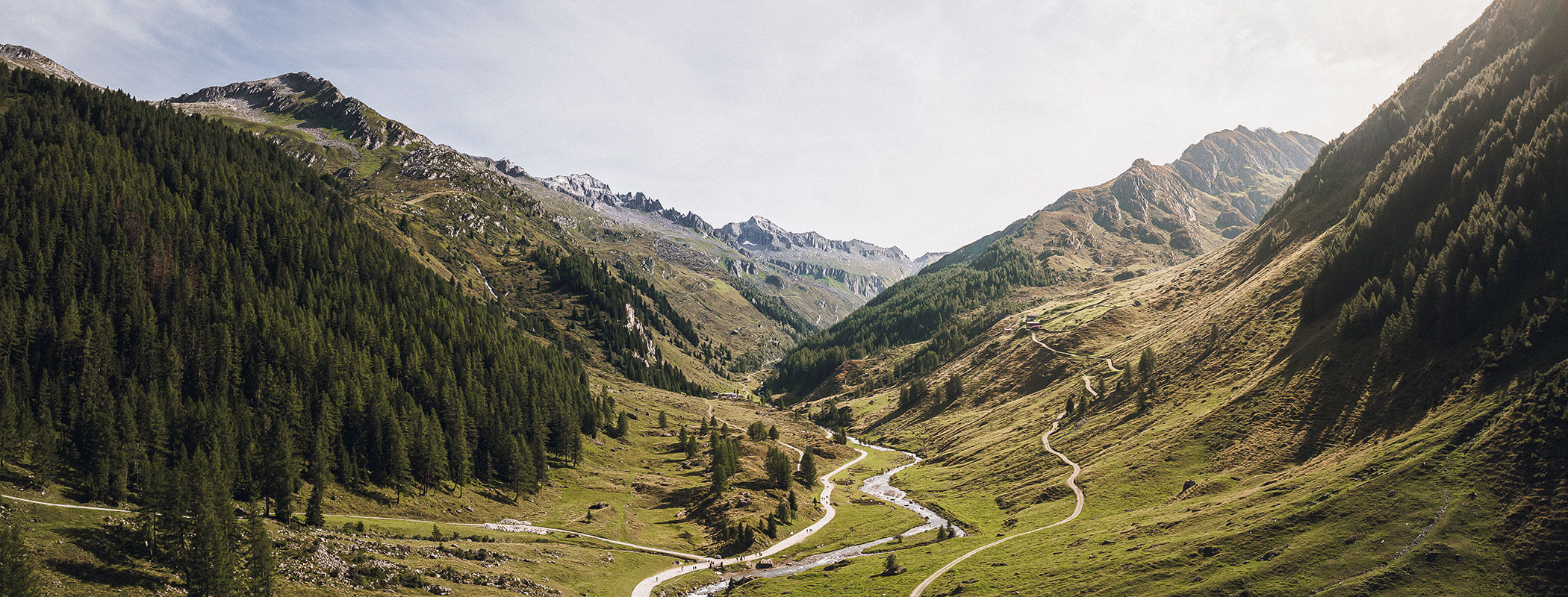 A winding road and river flow through a green mountain valley with trees under a partly cloudy sky. - Maurlechnhof