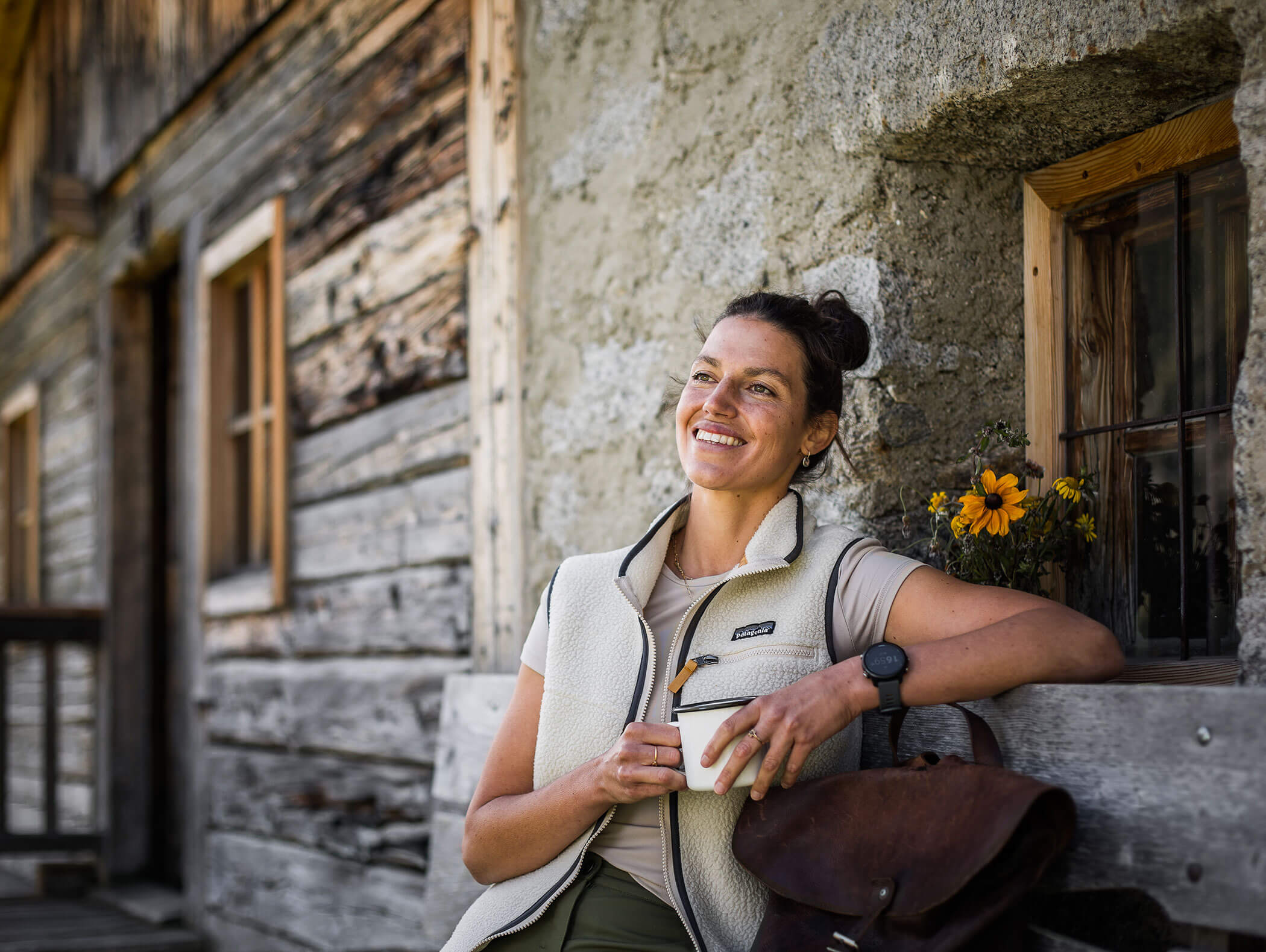 Smiling woman holding a cup, sitting on a bench by a rustic wooden house with flowers on the window sill. - Maurlechnhof