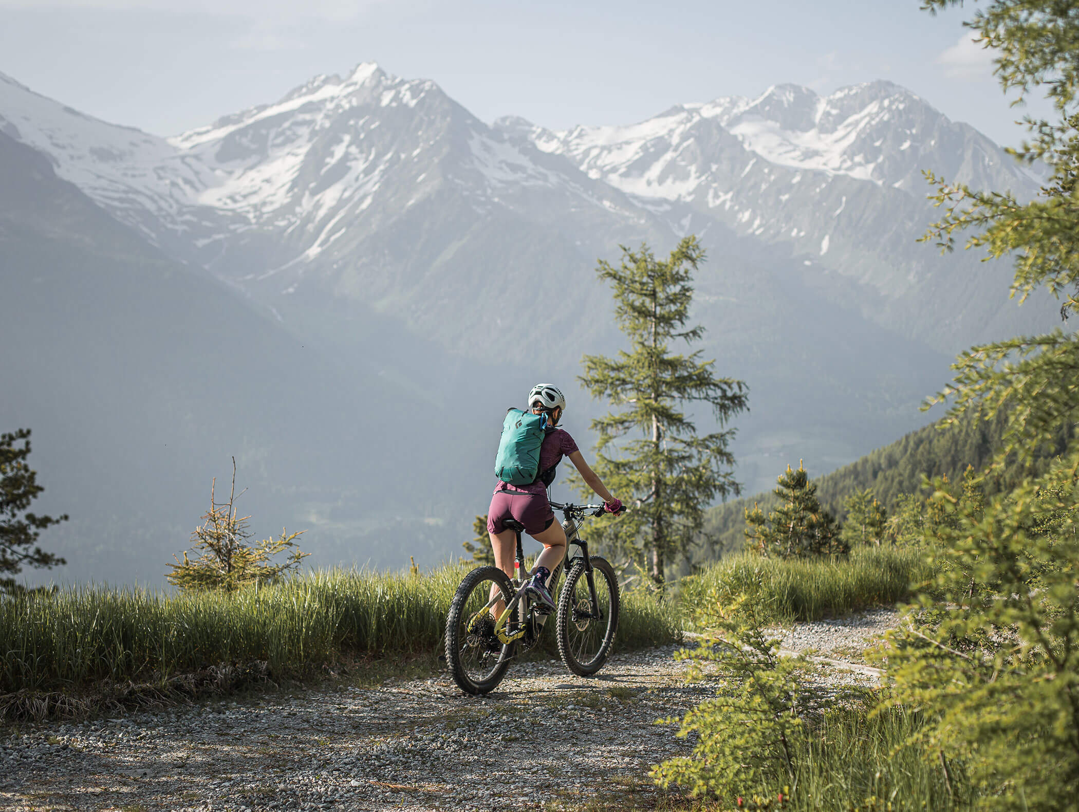 A cyclist rides a mountain trail with snow-capped peaks in the background on a sunny day. - Maurlechnhof