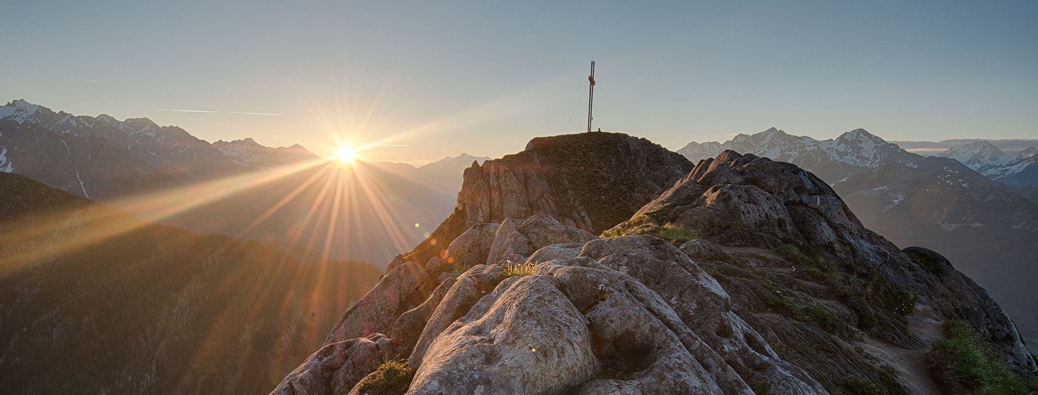 Sunrise over rocky mountain peak with a cross, surrounded by distant snow-capped mountains. - Maurlechnhof