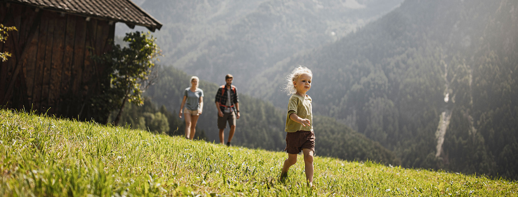 A young child walks ahead of two adults on a grassy hillside with mountains in the background. - Maurlechnhof