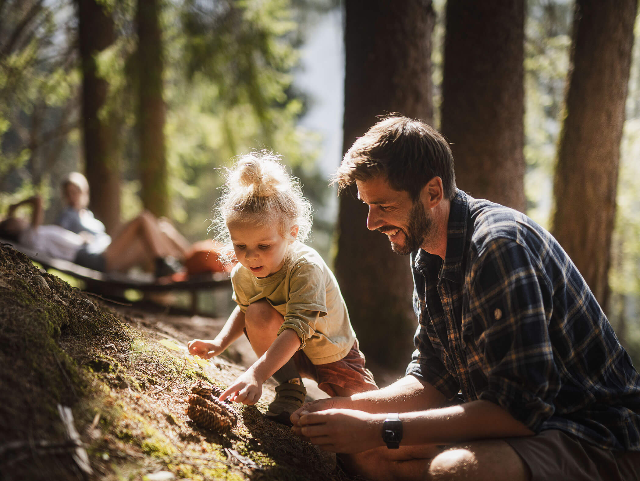 Man and young child exploring pinecones on a forest floor, with people relaxing in the background. - Maurlechnhof