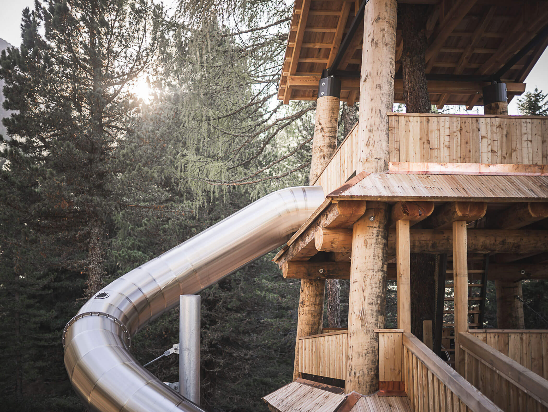 A wooden treehouse with a large metal slide winding down among tall pine trees in sunlight. - Maurlechnhof
