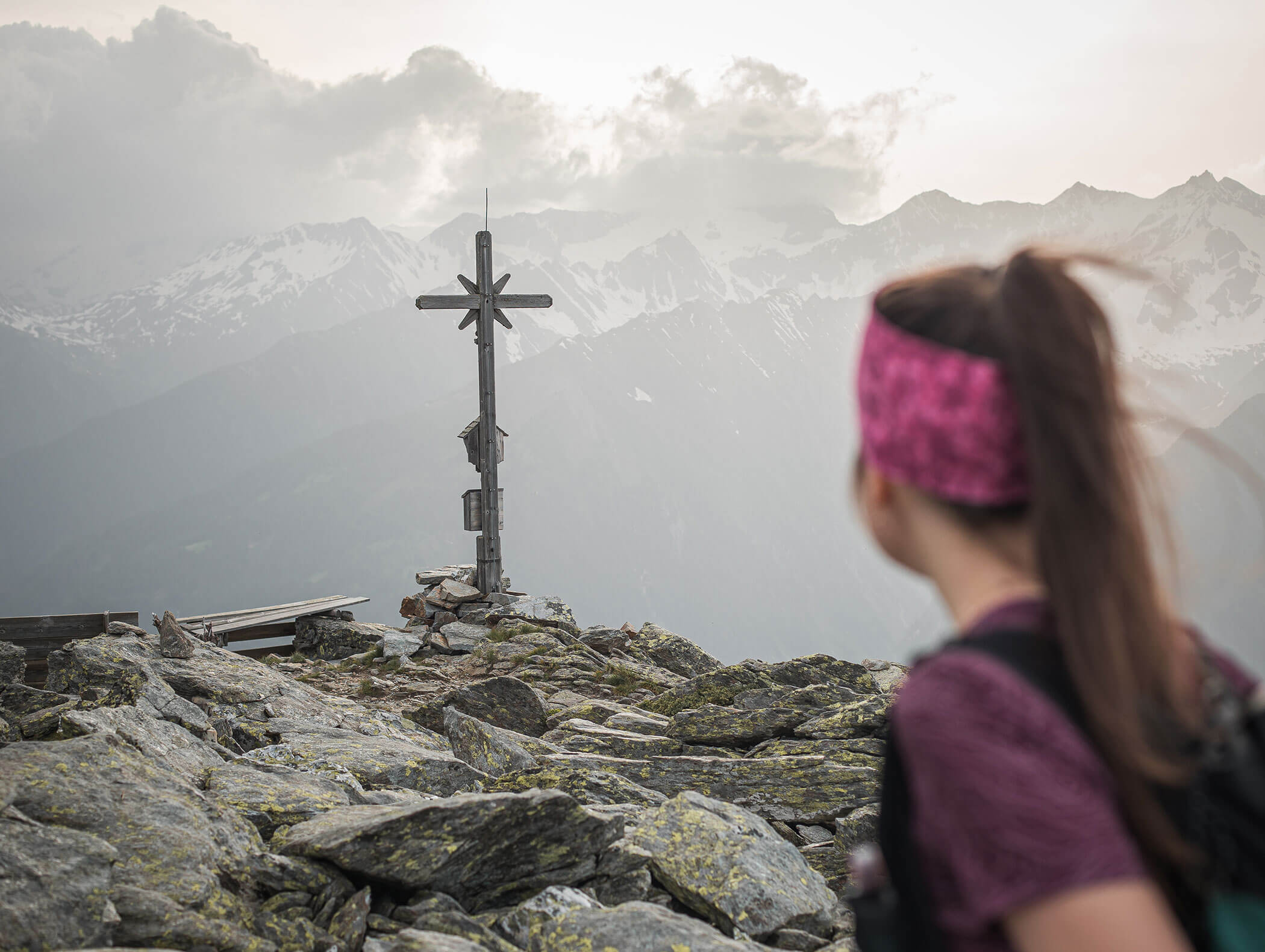 A woman looks at a summit cross on a rocky mountain peak with snowy mountains in the background. - Maurlechnhof