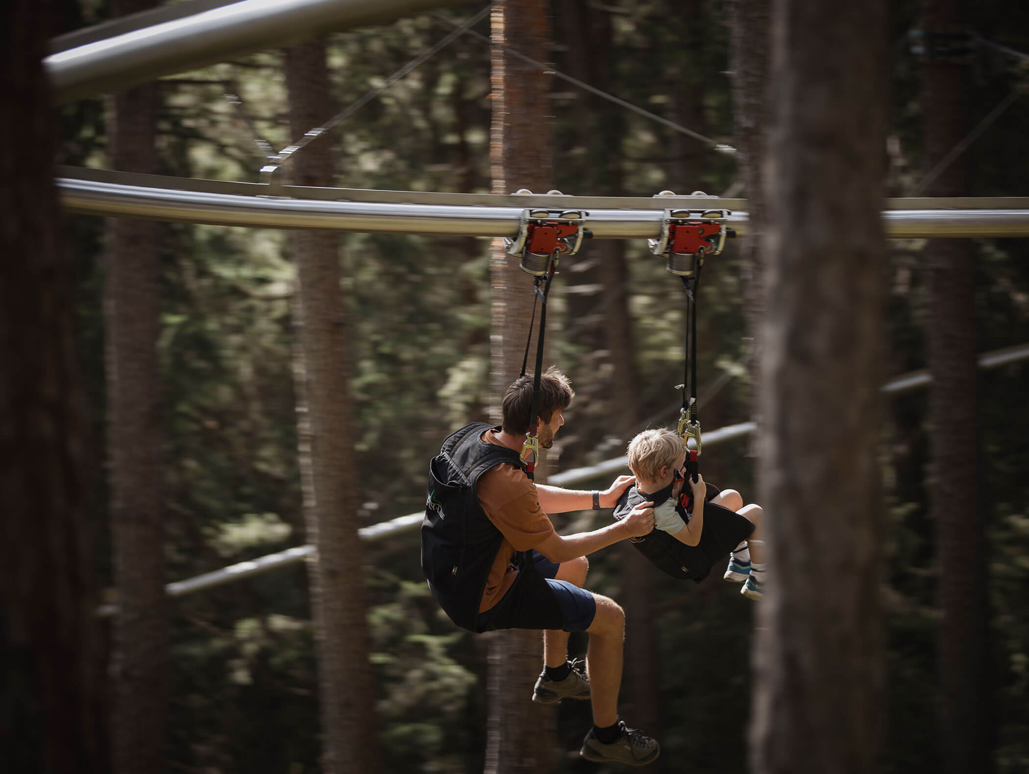 An adult and child wearing harnesses glide on a zipline track through a forest. - Maurlechnhof