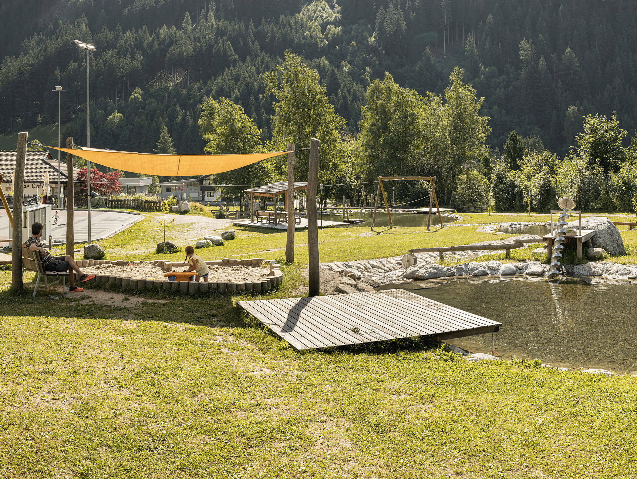 A child plays in a sandbox under a shade sail near a pond in a sunny park with swings and trees. - Maurlechnhof
