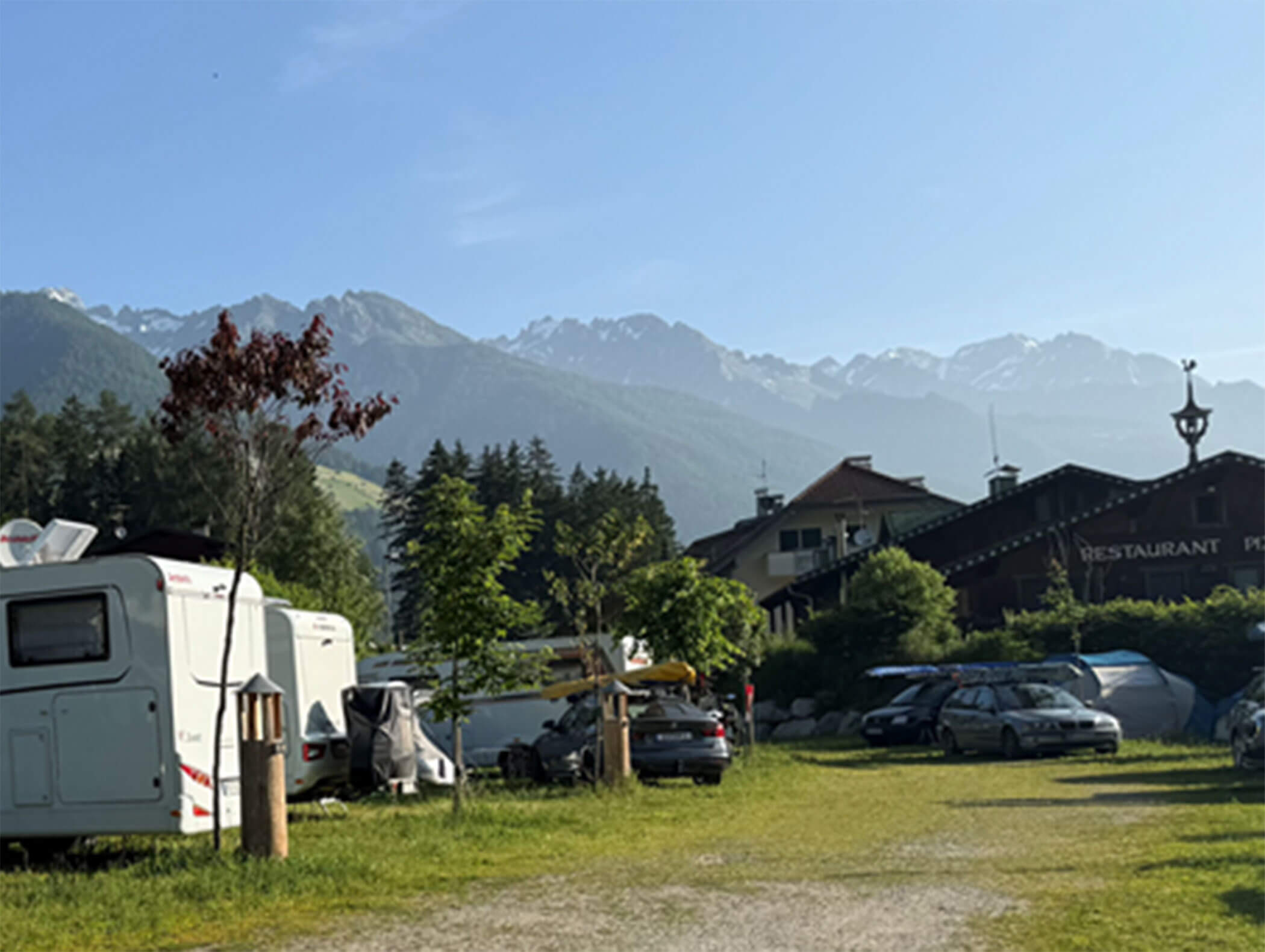 Campingplatz mit Wohnwagen und Autos, Berge im Hintergrund und ein Restaurantgebäude an einem sonnigen Tag. - Maurlechnhof