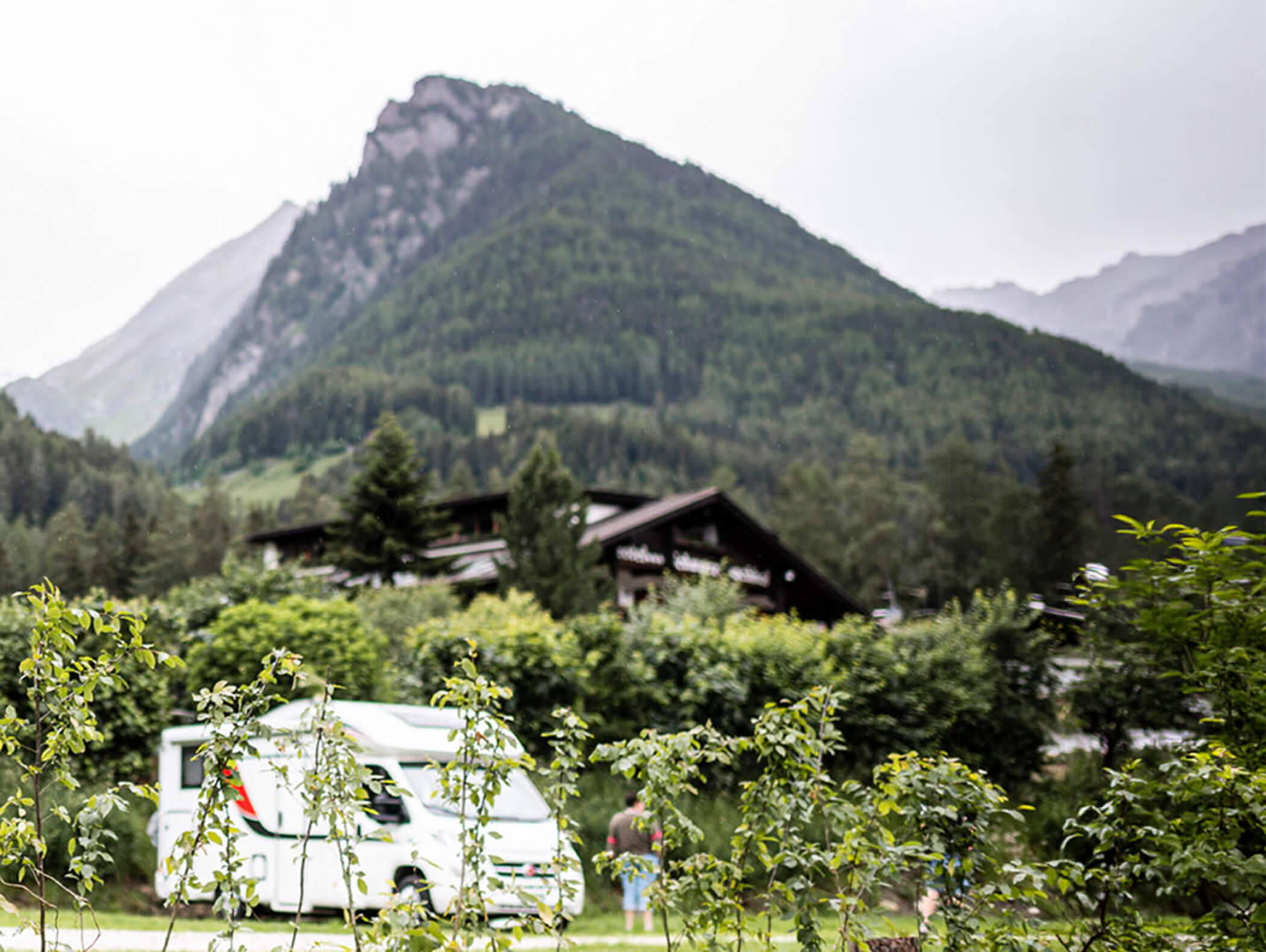 Ein weißes Wohnmobil parkt in der Nähe von grünen Büschen mit bewaldeten Bergen und einer Hütte im Hintergrund. - Maurlechnhof