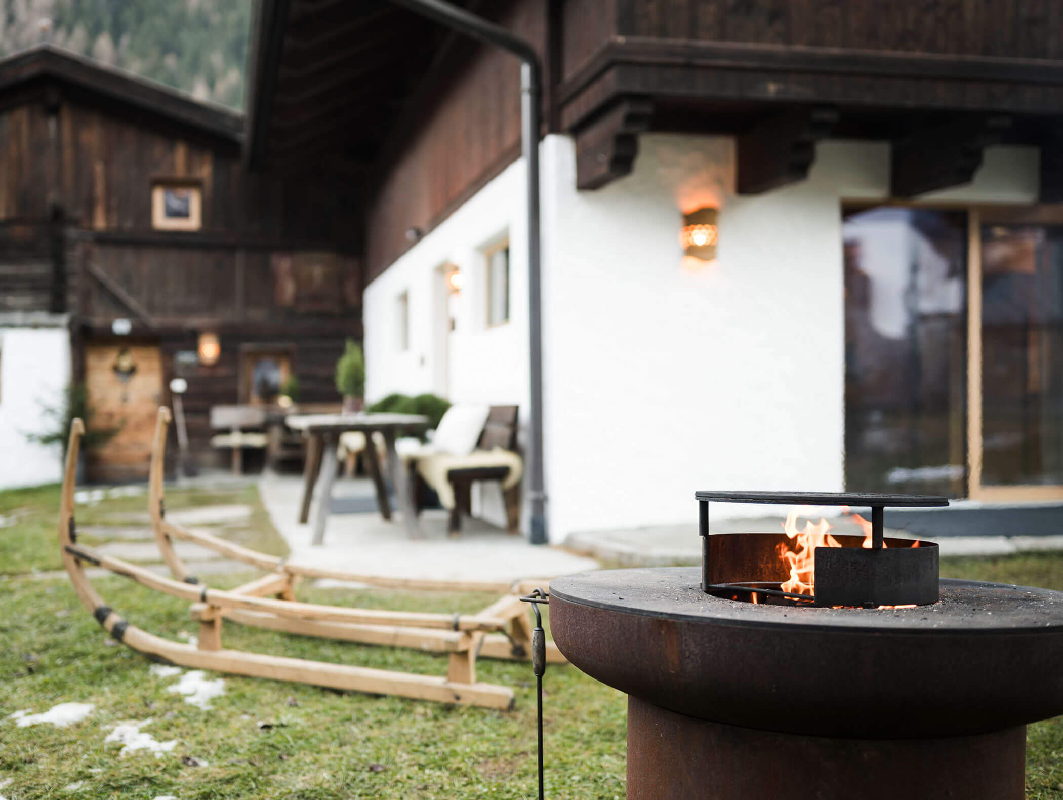 Eine Feuerstelle vor einer rustikalen Hütte mit Holzbänken und Tischen auf einer Wiese. - Maurlechnhof