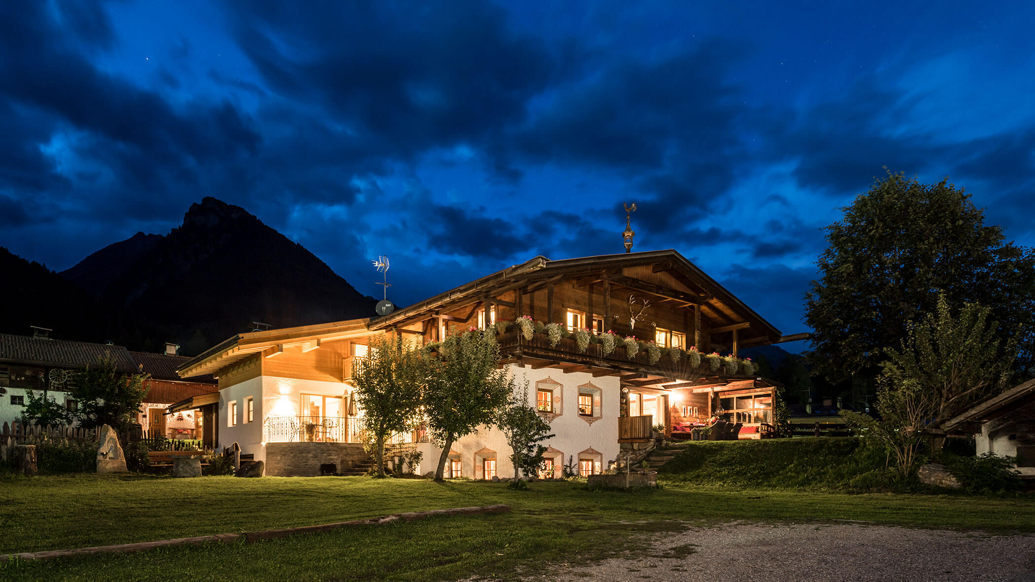 Ein warm beleuchtetes Alpenhaus in der Abenddämmerung, mit einem Berg und dramatischen Wolken im Hintergrund. - Maurlechnhof
