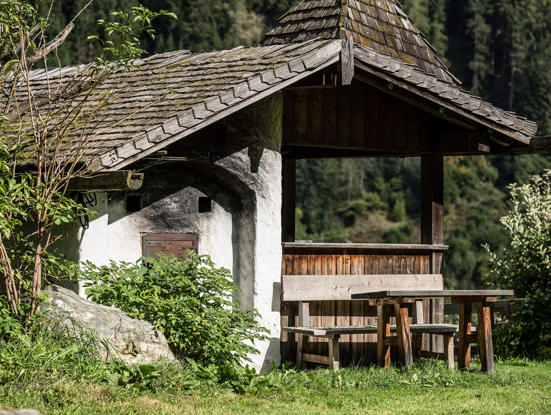 Hölzerner Picknicktisch unter einem rustikalen Unterstand mit Schindeldach in einem grasbewachsenen, bewaldeten Gebiet. - Maurlechnhof