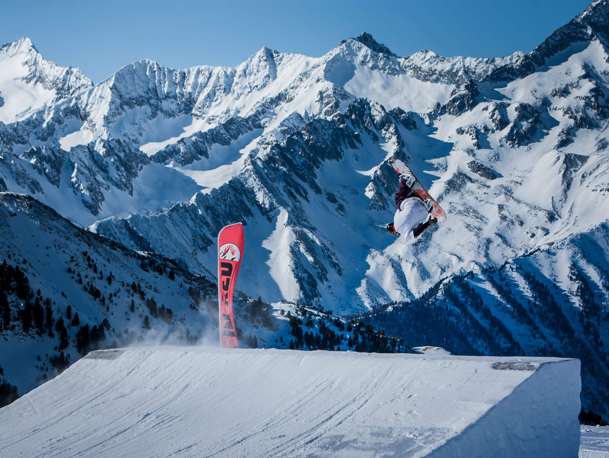 Ein Snowboarder springt von einer Schneerampe mit verschneiten Bergen und blauem Himmel im Hintergrund. - Maurlechnhof