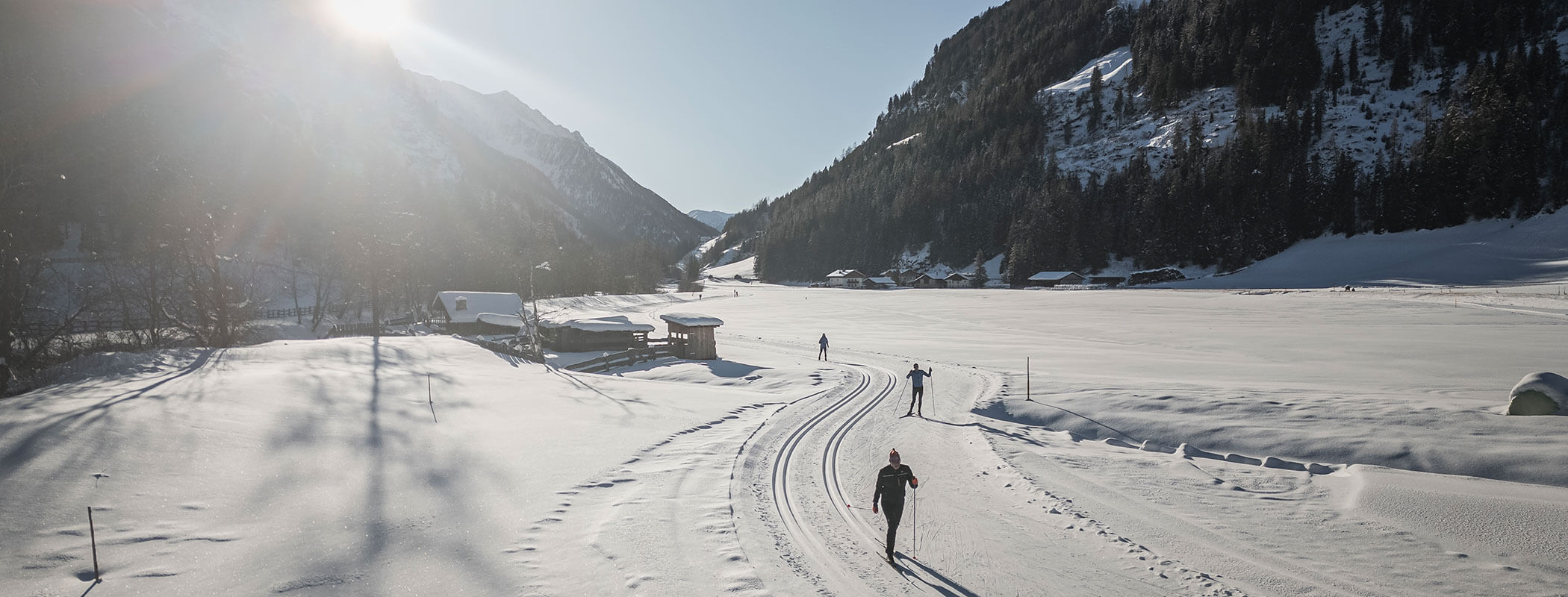 Drei Personen beim Skilanglauf auf verschneiten Loipen in einem Tal, umgeben von sonnenbeschienenen Bergen und Bäumen. - Maurlechnhof