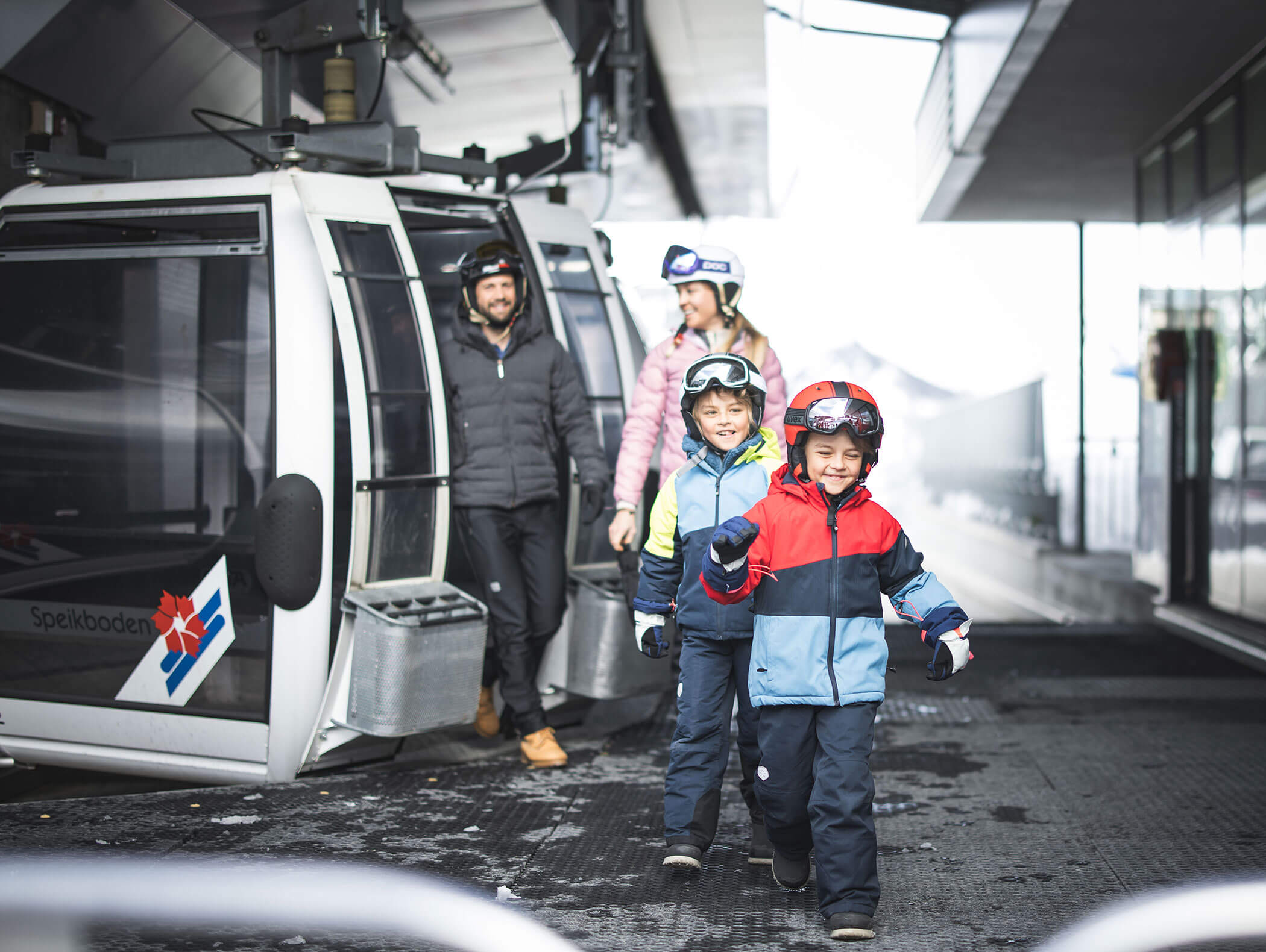 Zwei lächelnde Kinder in Skikleidung kommen aus einer Gondelbahn, gefolgt von zwei Erwachsenen in Winterkleidung und Helmen. - Maurlechnhof