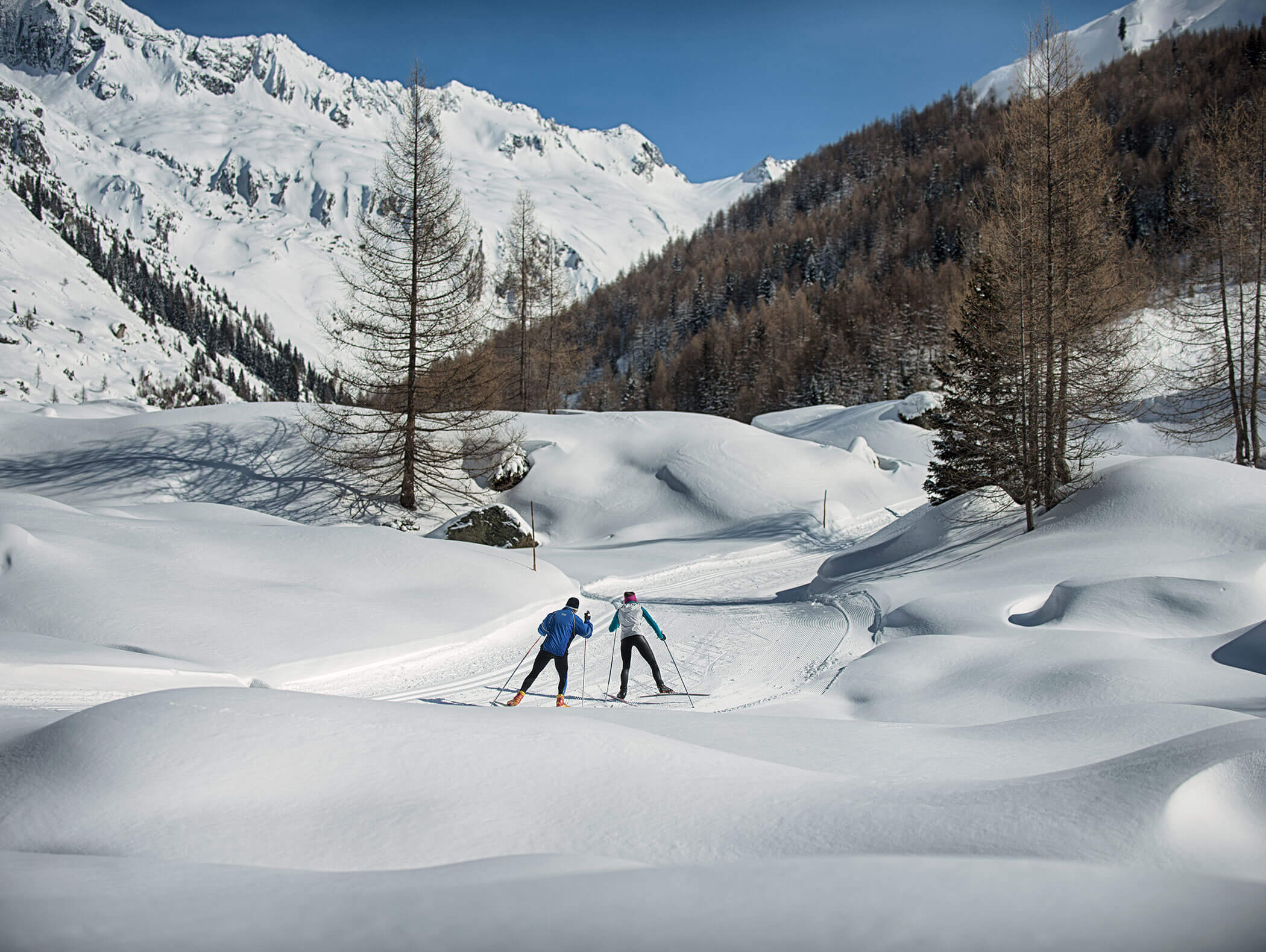 Zwei Personen beim Skilanglauf auf einer verschneiten Loipe, umgeben von Bergen und Kiefern. - Maurlechnhof