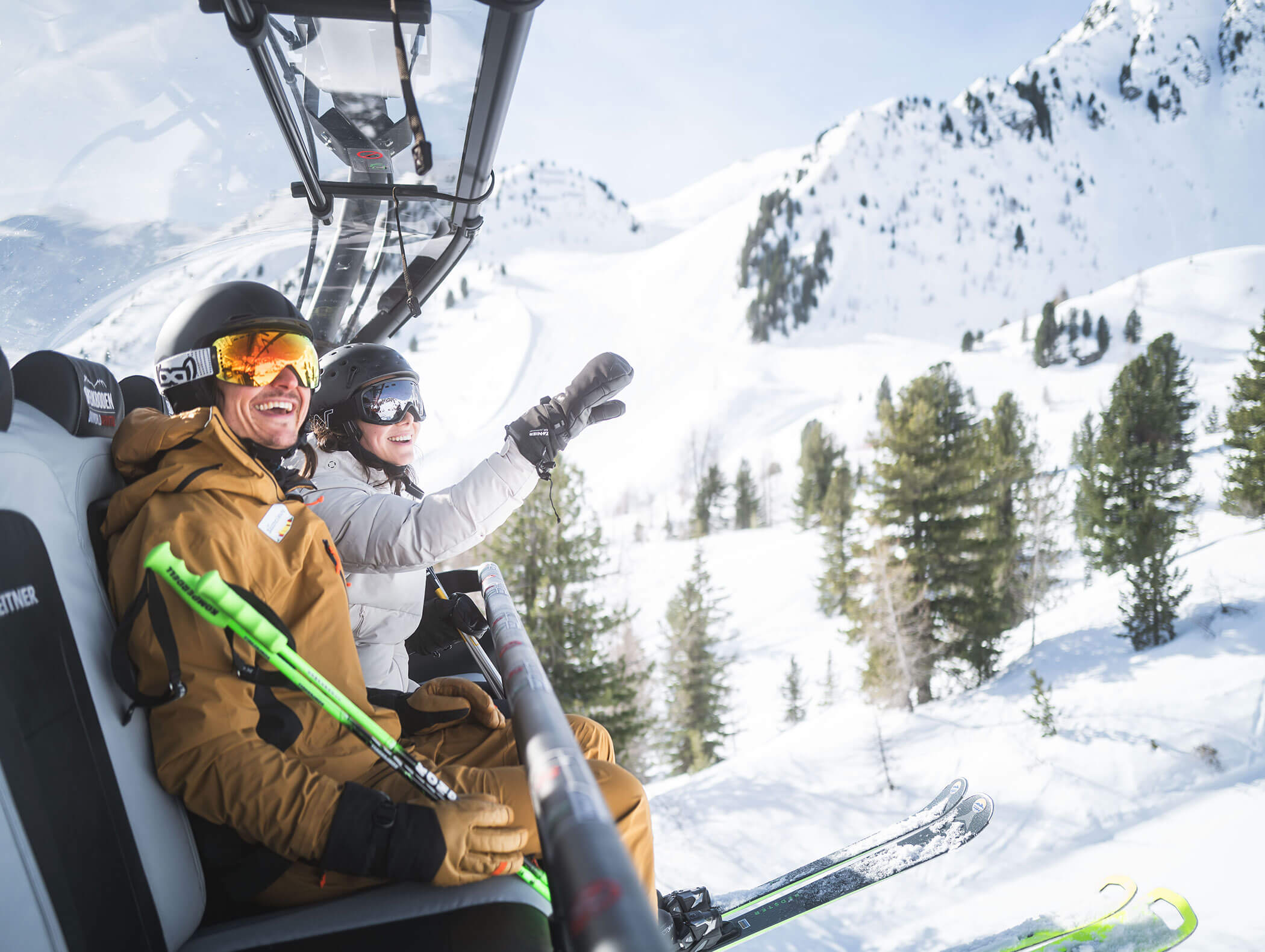 Zwei Skifahrer lächeln und winken, während sie mit einem Skilift über eine verschneite Berglandschaft fahren. - Maurlechnhof