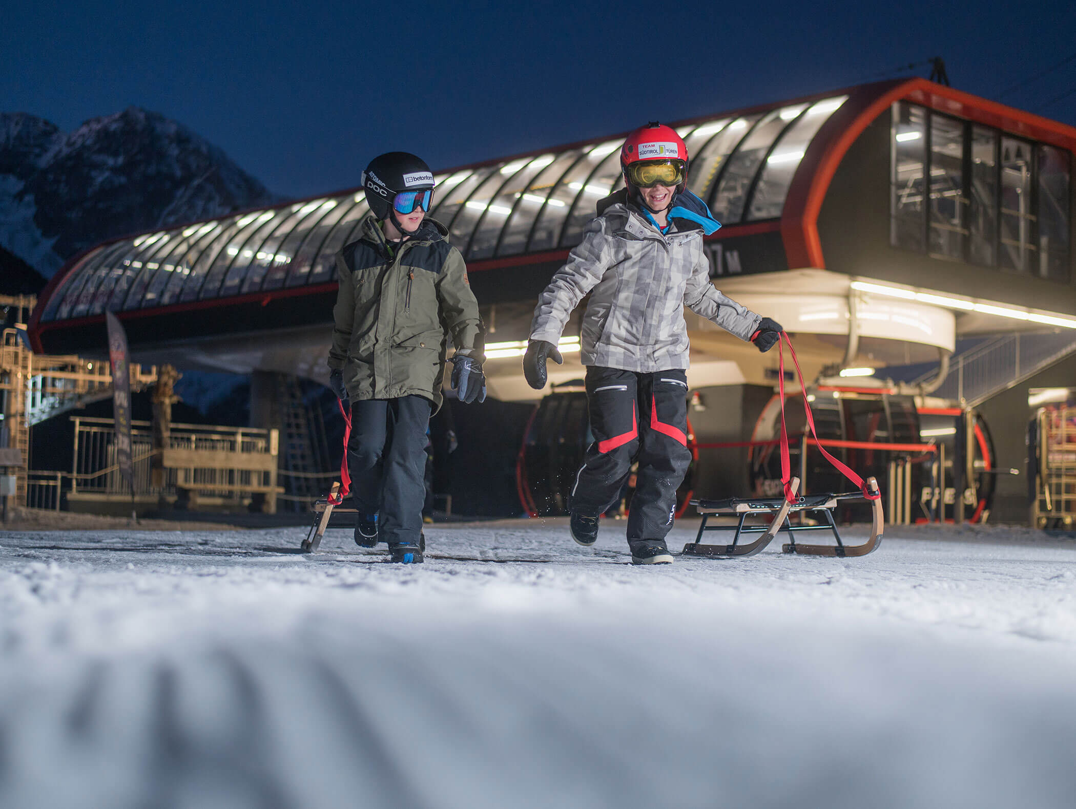 Zwei Kinder in Winterkleidung ziehen Schlitten auf Schnee in der Nähe eines Skilifts in der Abenddämmerung, mit Bergen im Hintergrund. - Maurlechnhof