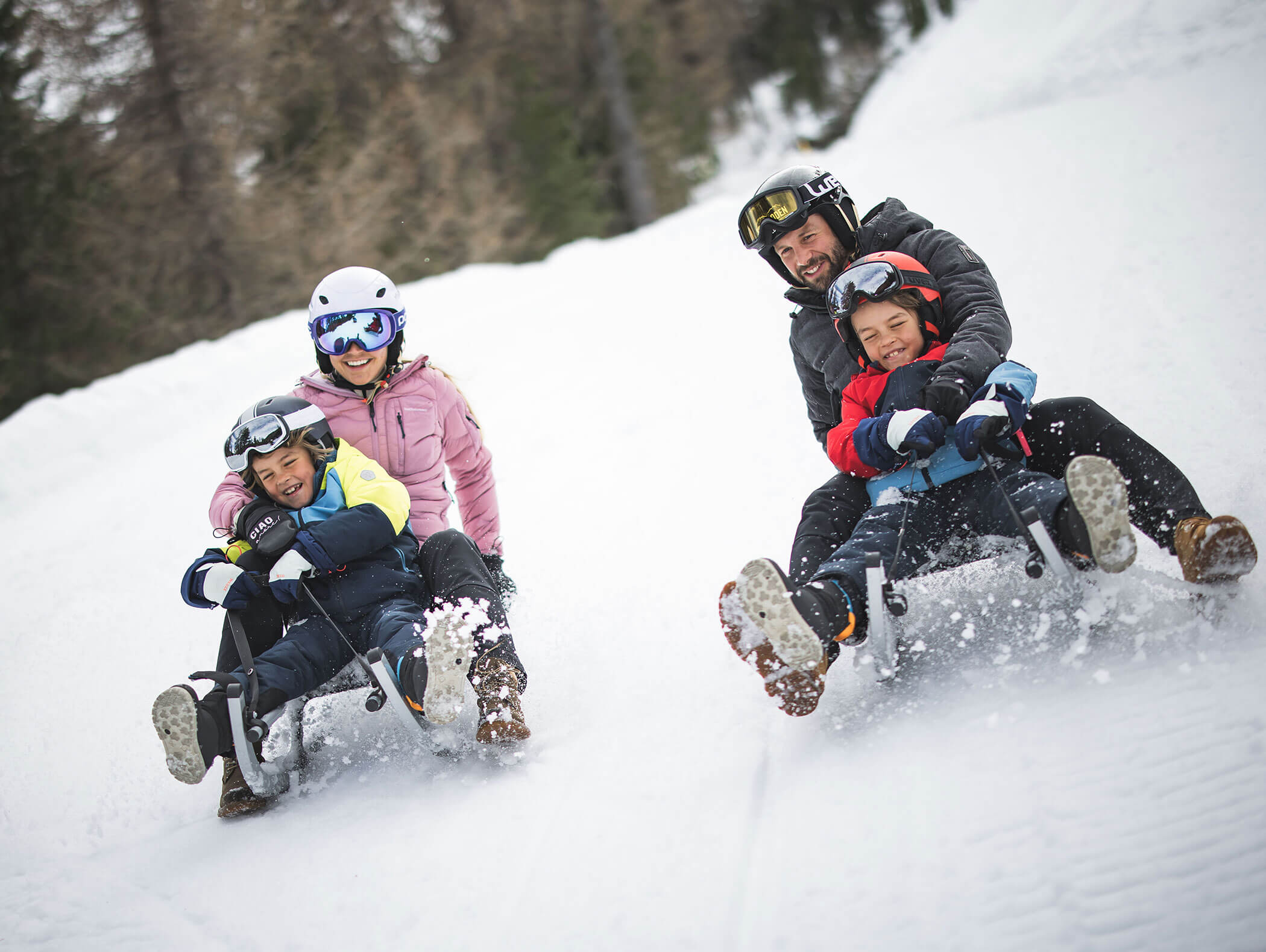 Eine vierköpfige Familie schlittelt einen verschneiten Hügel hinunter, lächelt und genießt einen gemeinsamen Wintertag. - Maurlechnhof