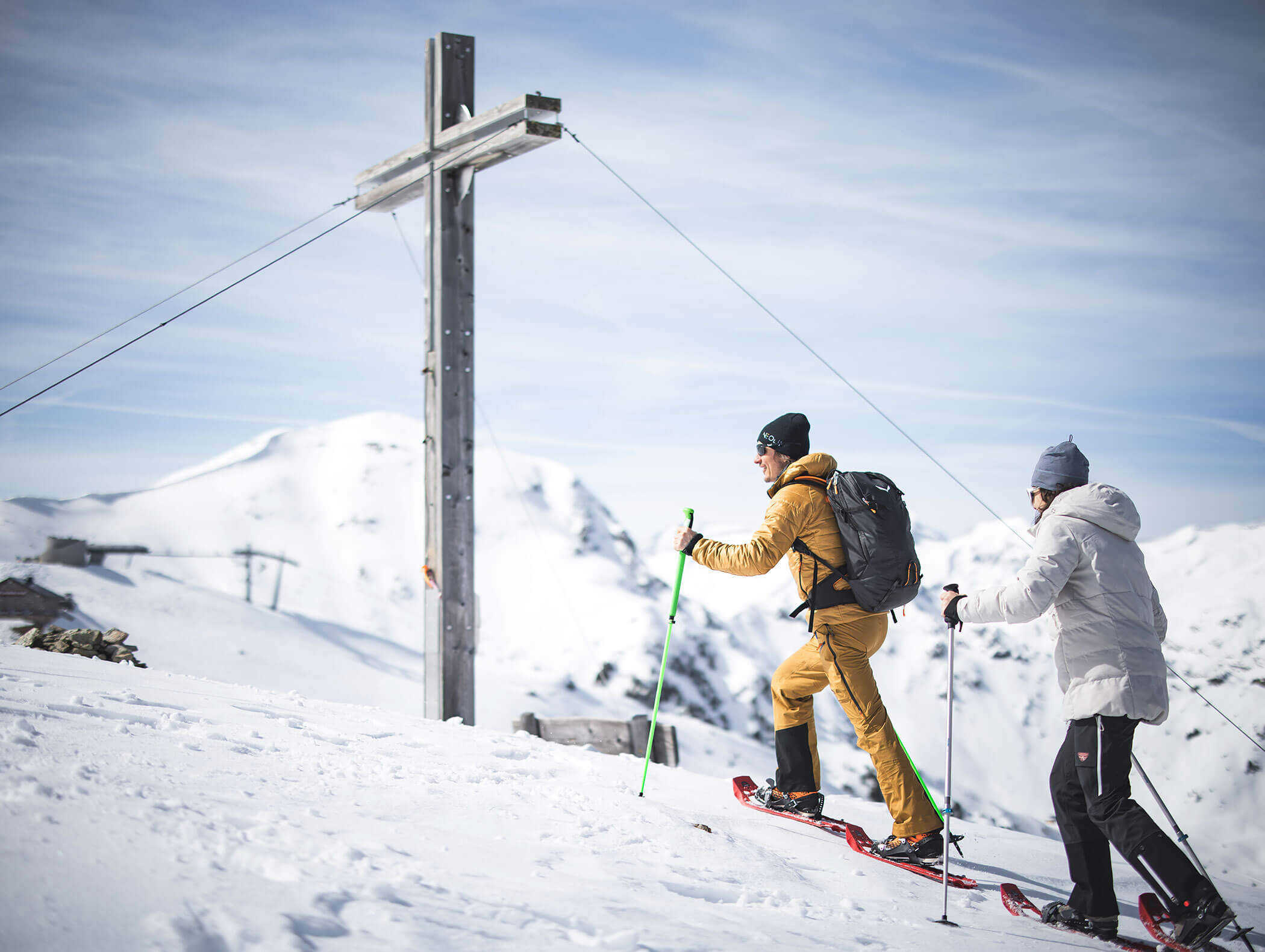 Zwei Personen in Winterausrüstung wandern an einem sonnigen Tag einen verschneiten Berghang in der Nähe eines großen Holzkreuzes hinauf. - Maurlechnhof