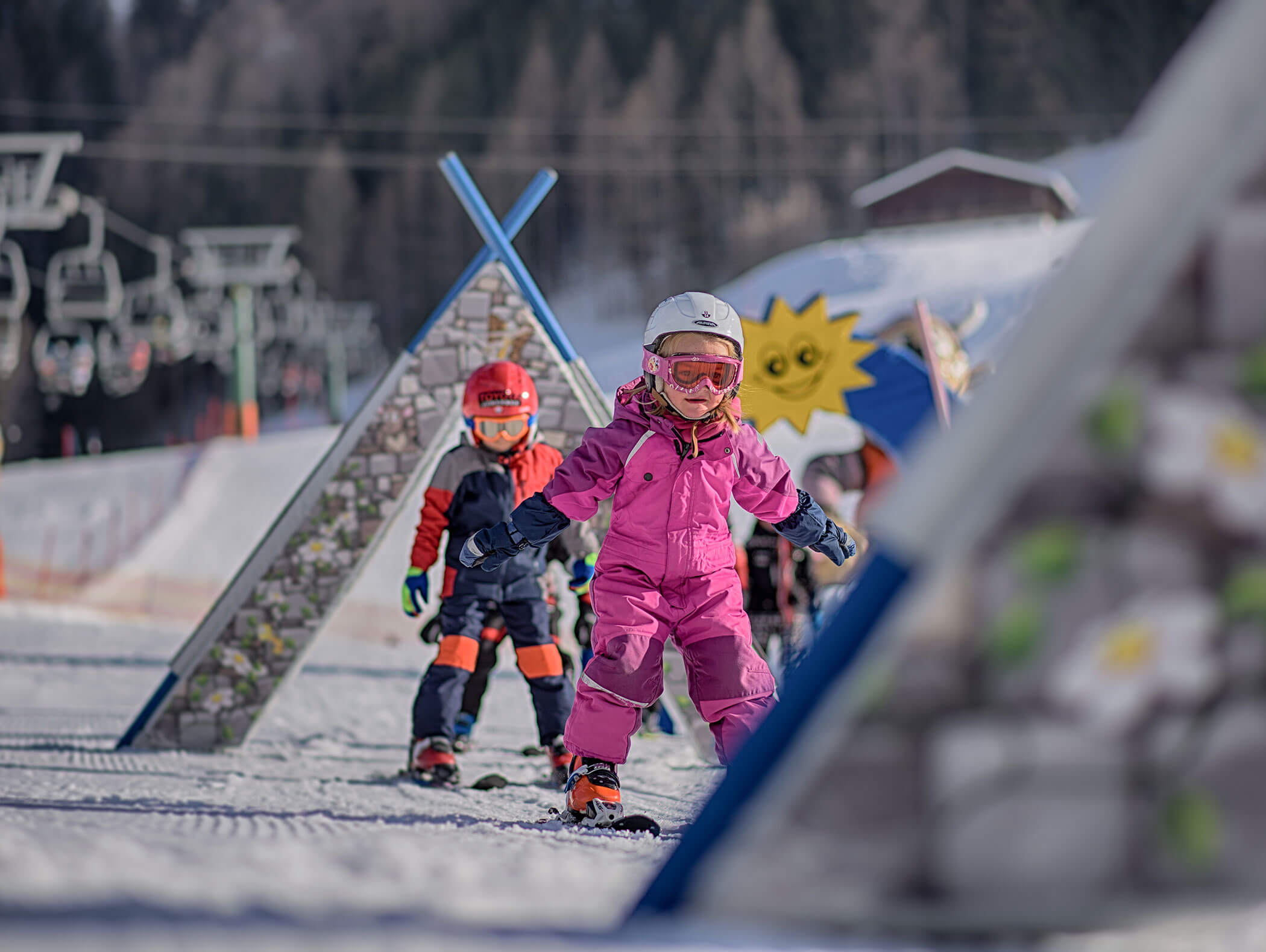 Junge Kinder fahren auf einer verschneiten Piste Ski, tragen Helme und bunte Skianzüge und sind von lustigen Hindernissen umgeben. - Maurlechnhof