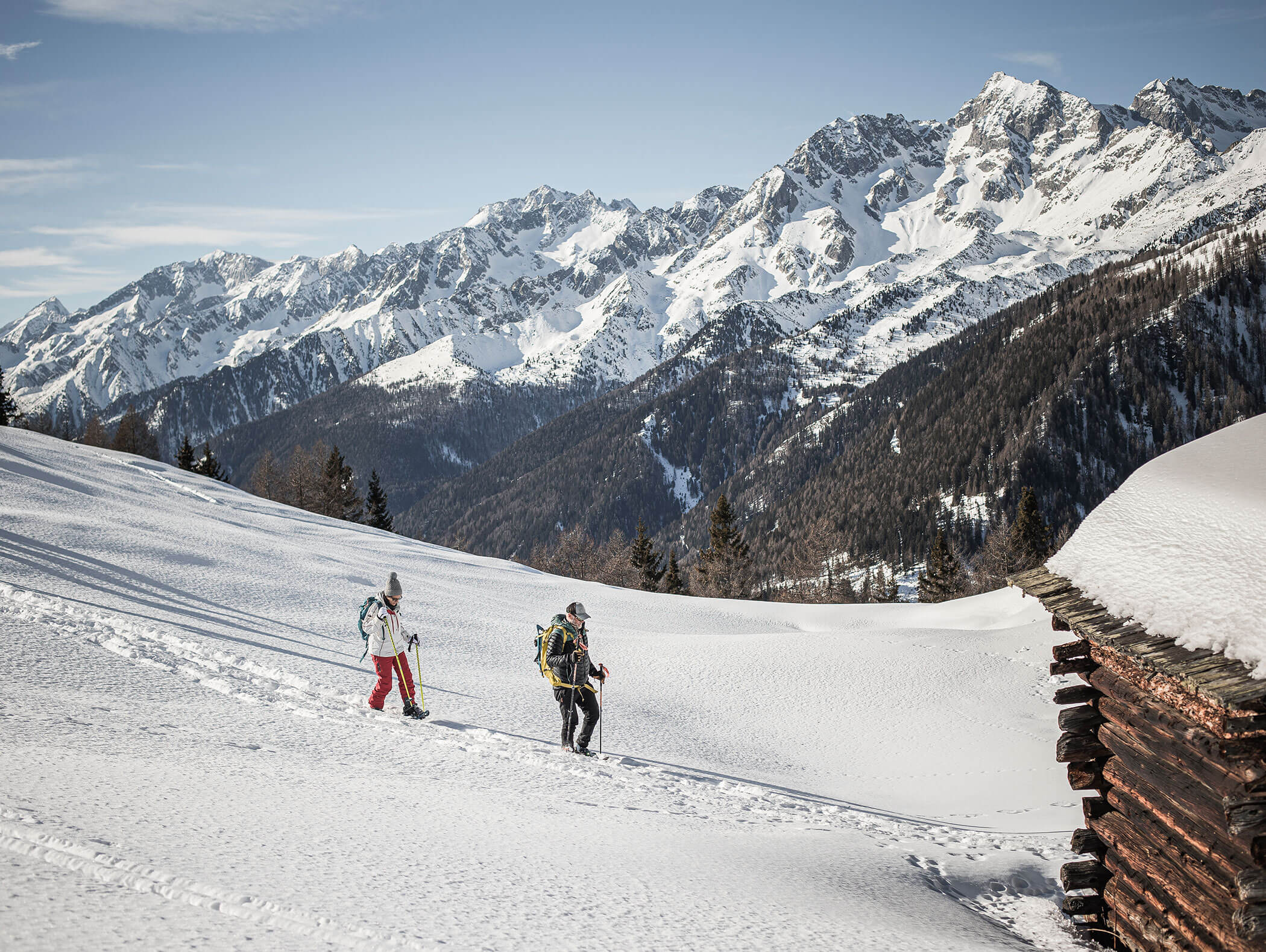 Zwei Personen wandern mit Rucksäcken einen verschneiten Berghang hinauf, umgeben von alpinen Gipfeln und Tannenbäumen. - Maurlechnhof